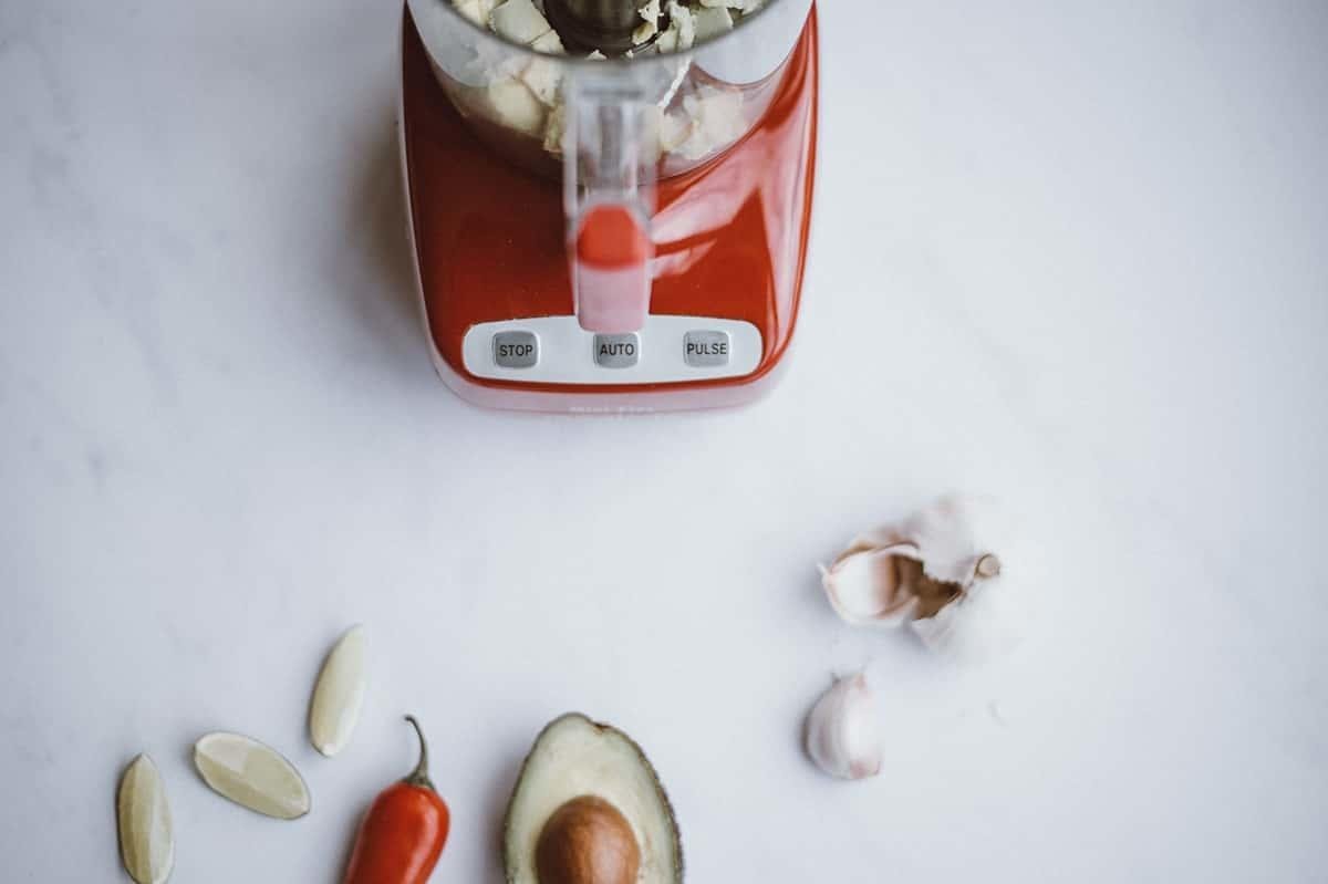 A modern food processor surrounded by various nuts on a clean marble countertop.